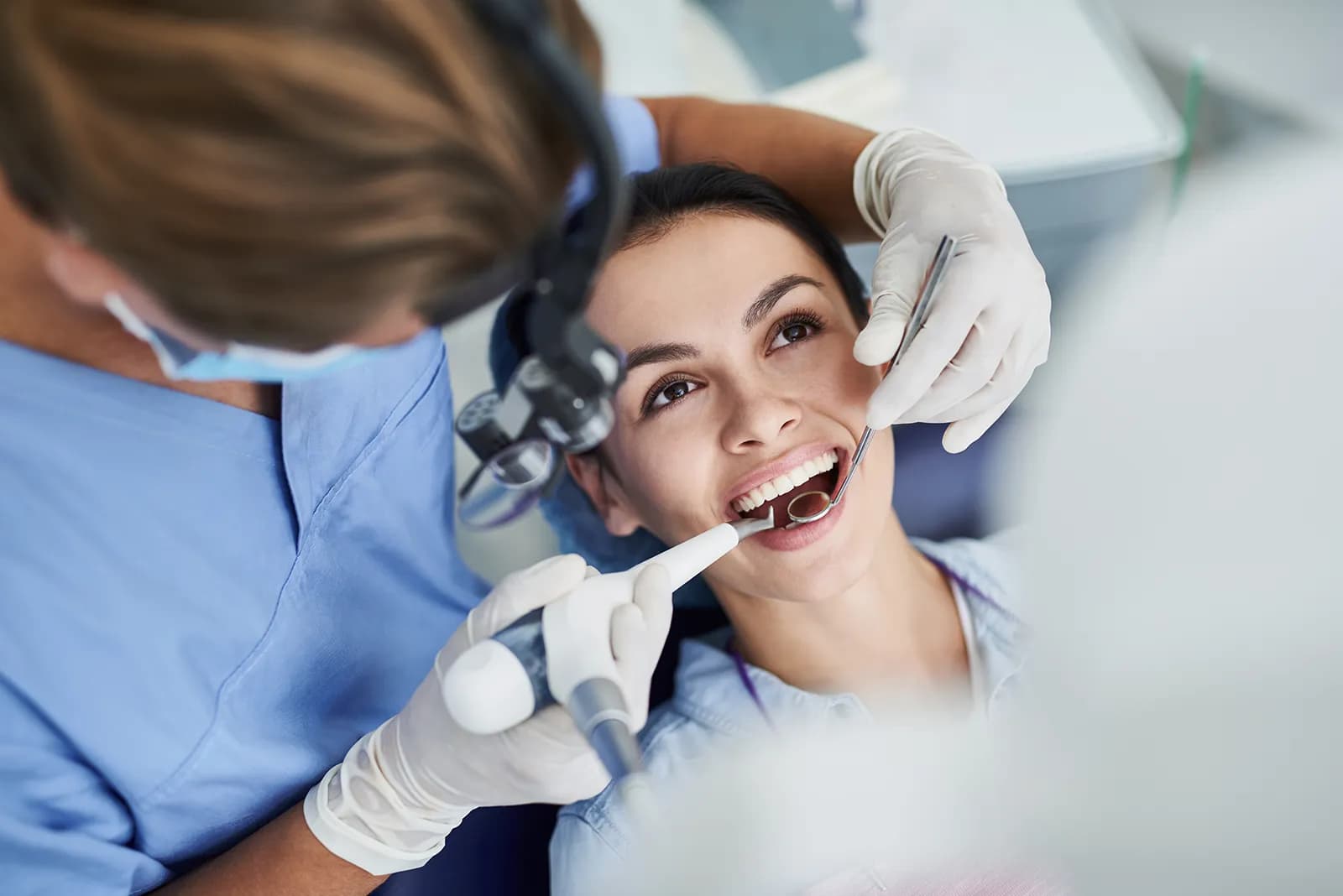 A dental hygienist in blue scrubs and magnifying loupes cleaning the teeth of a smiling patient in a bright treatment room.